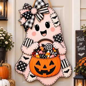 May include: A light pink ghost-shaped door hanger featuring a large black and white buffalo plaid bow, patches with bat and polka dot patterns, and a bright orange jack-o'-lantern bucket filled with various candies, suspended by a thick rope.