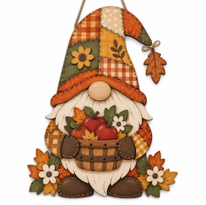 Puede incluir: Un adorno de pared de madera con forma de gnomo que luce un sombrero de patchwork en tonos naranja, verde y crema. El gnomo sostiene una cesta de manzanas rojas y hojas otoñales, con flores y hojas en la base y un colgante en forma de hoja.