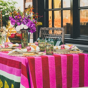 Op de afbeelding: Een roze en groene gestreepte tafelkleed met een geel rozenpatroon aan de rand. Het tafelkleed is gedekt voor een maaltijd met borden, glazen en bestek.
