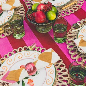 Op de afbeelding: Een tafeldekking met roze en rode gestreepte tafelkleed, geweven placemats, witte borden met gouden rand, groene glazen en een kom met fruit. Servetten zijn gevouwen en vastgebonden met een roze en witte servetring.