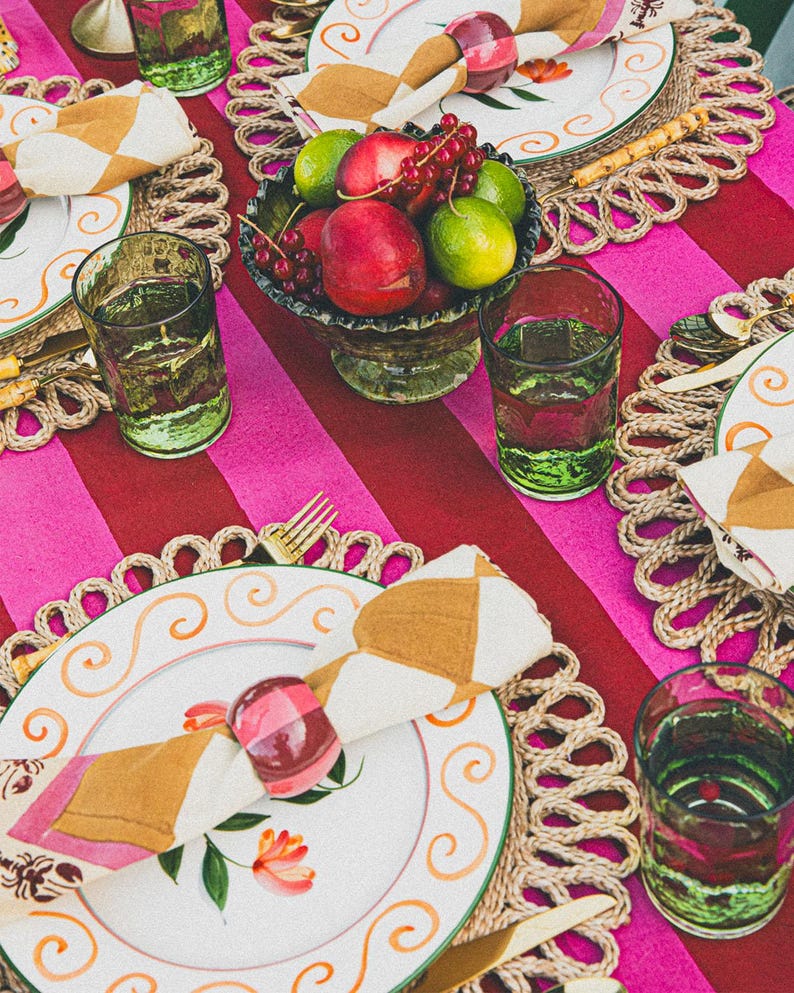 Op de afbeelding: Een tafeldekking met roze en rode gestreepte tafelkleed, geweven placemats, witte borden met gouden rand, groene glazen en een kom met fruit. Servetten zijn gevouwen en vastgebonden met een roze en witte servetring.