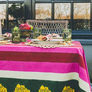 Op de afbeelding: Een gestreept tafelkleed in roze, wit, groen en bordeauxrood met gele rozenmotieven. Het tafelkleed is gedekt met een bord, een servet en een glas.
