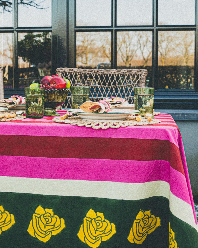 Op de afbeelding: Een gestreept tafelkleed in roze, wit, groen en bordeauxrood met gele rozenmotieven. Het tafelkleed is gedekt met een bord, een servet en een glas.