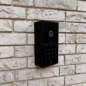 Può includere: Un tastierino nero con una telecamera e un tastierino numerico. Il tastierino è montato su un muro di mattoni bianchi.