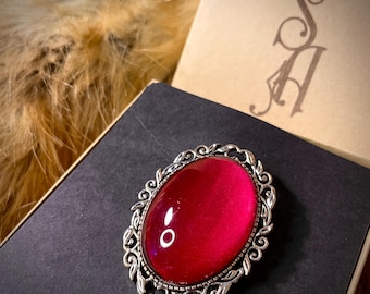 May include: A red glass cabochon set in a silver ornate frame. The cabochon has a slight iridescent quality.