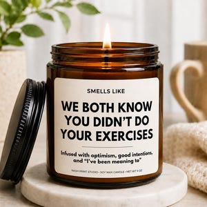 May include: A brown glass jar candle with a black lid and a white label that reads: SMELLS LIKE WE BOTH KNOW YOU DIDN'T DO YOUR EXERCISES. Infused with optimism, good intentions, and “I’ve been meaning to”. NASH PRINT STUDIO SOY WAX CANDLE NET WT 9 OZ.