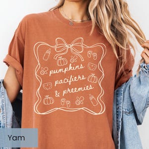 May include: A terracotta-coloured t-shirt with a white graphic that reads "pumpkins, pacifiers, & preemies" inside a decorative frame. The design includes illustrations of pumpkins, baby bottles, pacifiers, and nappies. The shirt has rolled-up sleeves.