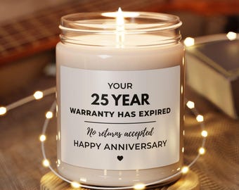 May include: A lit candle in a glass jar with a white label that reads "YOUR 25 YEAR WARRANTY HAS EXPIRED No returns accepted HAPPY ANNIVERSARY" on a wooden coaster. A string of lights and a guitar are in the background.