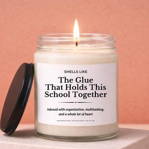 May include: A lit candle in a clear glass jar with a black lid. The label reads "SMELLS LIKE The Glue That Holds This School Together." The candle sits on a square, light-colored block against a peach background.