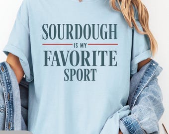 Könnte beinhalten: Hellblaues T-Shirt mit dem Text "SOURDOUGH IS MY FAVORITE SPORT" in einem zweizeiligen Design. Das Shirt besteht aus einem weichen Material, ideal für den Alltag. Eine Jeansjacke ist über den Schultern getragen.