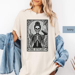 May include: Ivory t-shirt with a black and white graphic of a skeleton hairstylist holding scissors and a hairdryer. The graphic is framed with floral elements and the text "THE STYLIST". The shirt is a casual, unisex style.