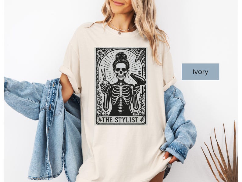 May include: Ivory t-shirt with a black and white graphic of a skeleton hairstylist holding scissors and a hairdryer. The graphic is framed with floral elements and the text "THE STYLIST". The shirt is a casual, unisex style.