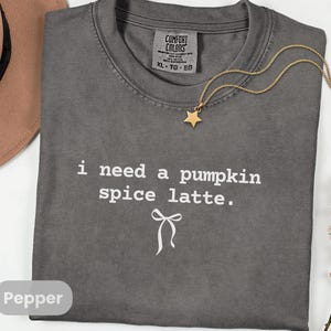 May include: A gray Comfort Colors t-shirt with the text "i need a pumpkin spice latte." A gold star pendant necklace and a brown hat are also visible. The shirt is size XL-TG-EG.