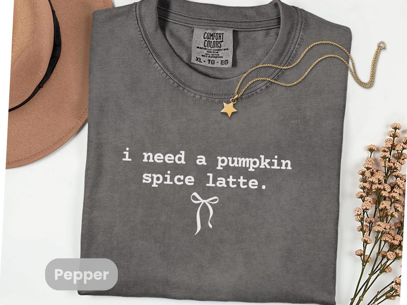 May include: A gray Comfort Colors t-shirt with the text "i need a pumpkin spice latte." A gold star pendant necklace and a brown hat are also visible. The shirt is size XL-TG-EG.