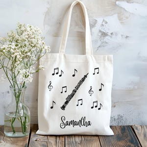May include: Cream-colored tote bag featuring a black oboe, musical notes, and the name "Samantha." The bag has a simple design with a handle for carrying. A clear glass vase with white flowers sits to the left.