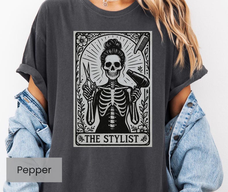 May include: Dark grey t-shirt with a black and white graphic of a skeleton hairstylist holding scissors and a hairdryer. The graphic includes a comb and the text "THE STYLIST" in a decorative frame. The shirt is a Pepper colour.