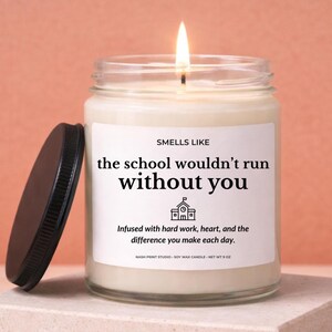 Puede incluir: Una vela de cera de soja encendida en un tarro de cristal con tapa negra. La etiqueta dice "Smells Like the school wouldn't run without you". La vela está impregnada de trabajo duro, corazón y la diferencia que haces cada día.