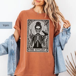 May include: A terracotta-coloured t-shirt with a black and white graphic of a skeleton hairstylist holding scissors and a hairdryer. The text "THE STYLIST" is below the image. The shirt is a casual style.