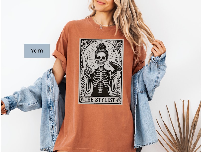 May include: A terracotta-coloured t-shirt with a black and white graphic of a skeleton hairstylist holding scissors and a hairdryer. The text "THE STYLIST" is below the image. The shirt is a casual style.