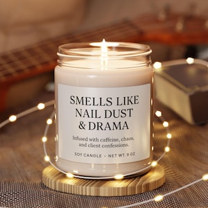 May include: A lit soy candle in a glass jar with the text "SMELLS LIKE NAIL DUST & DRAMA." The candle is on a wooden coaster and surrounded by string lights. The candle weighs approximately 255 grams.