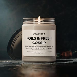 Puede incluir: Una vela de cera de soja encendida en un frasco de vidrio transparente con tapa negra. La etiqueta dice "SMELLS LIKE FOILS & FRESH GOSSIP". La vela pesa 255 gramos y tiene el texto "Infused with root touch-ups, rumor updates, and the kind of gossip that never leaves the chair."