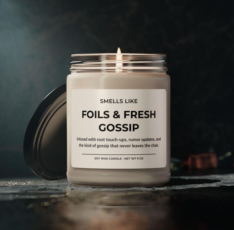 Puede incluir: Una vela de cera de soja encendida en un frasco de vidrio transparente con tapa negra. La etiqueta dice "SMELLS LIKE FOILS & FRESH GOSSIP". La vela pesa 255 gramos y tiene el texto "Infused with root touch-ups, rumor updates, and the kind of gossip that never leaves the chair."