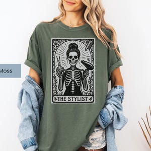 May include: A moss green t-shirt with a black and white graphic of a skeleton in a stylist pose, holding scissors and a hairdryer. The graphic is framed with floral elements and the text "THE STYLIST".
