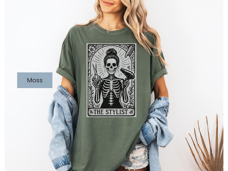 May include: A moss green t-shirt with a black and white graphic of a skeleton in a stylist pose, holding scissors and a hairdryer. The graphic is framed with floral elements and the text "THE STYLIST".