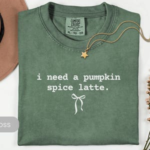 May include: A moss green t-shirt with the text "i need a pumpkin spice latte." A gold necklace with a star pendant rests on the shirt. The shirt is folded, and a brown hat and dried flowers are in the background.