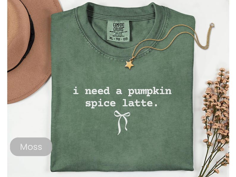 May include: A moss green t-shirt with the text "i need a pumpkin spice latte." A gold necklace with a star pendant rests on the shirt. The shirt is folded, and a brown hat and dried flowers are in the background.