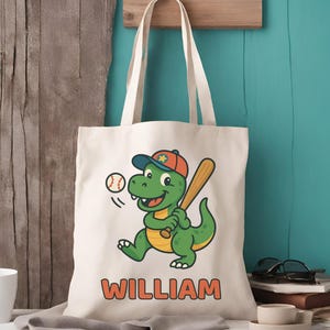 May include: Cream-colored tote bag featuring a cartoon dinosaur in a baseball cap, holding a bat and ready to hit a baseball. The name "WILLIAM" is printed in orange block letters below the dinosaur. The bag has cream-colored handles.