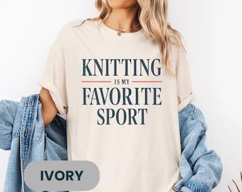 Op de afbeelding: Ivoor kleurig t-shirt met de tekst "KNITTING IS MY FAVORITE SPORT" in marineblauwe letters. De tekst is gecentreerd, met een rode lijn die de woorden scheidt. Het shirt heeft een relaxte pasvorm en opgerolde mouwen.