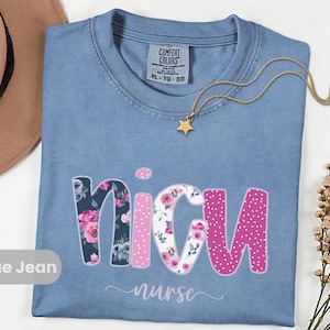 May include: Blue jean t-shirt with the word "NICU" in a floral and polka dot design. The word "nurse" is written below in a script font. A gold star pendant necklace rests on the shirt.