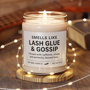 May include: A lit soy wax candle in a glass jar with a white label that reads "Smells Like Lash Glue & Gossip." The candle sits on a wooden coaster and is surrounded by string lights. Net weight is 255 grams.