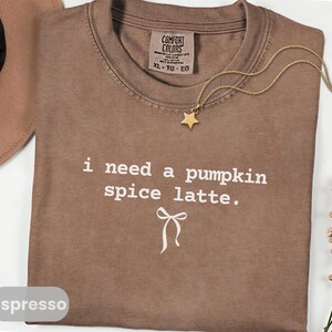 May include: Espresso-colored t-shirt with the text "i need a pumpkin spice latte." A gold-colored star pendant hangs from a delicate chain. A brown hat and a sprig of flowers are in the background. The shirt is size XL.
