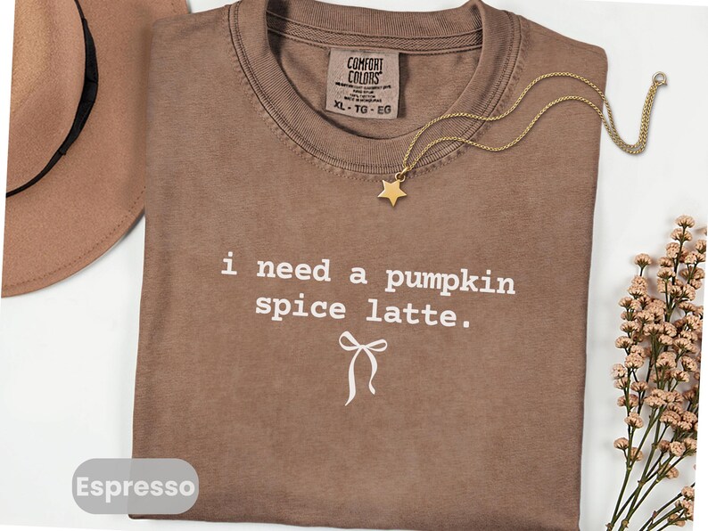 May include: Espresso-colored t-shirt with the text "i need a pumpkin spice latte." A gold-colored star pendant hangs from a delicate chain. A brown hat and a sprig of flowers are in the background. The shirt is size XL.