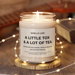 May include: A lit soy wax candle in a glass jar with the text "Smells Like A Little Tox & A Lot of Tea." The candle is on a wooden coaster and surrounded by string lights. The candle weighs approximately 255 grams.
