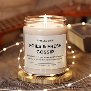 Puede incluir: Una vela de cera de soja encendida en un tarro de cristal con una etiqueta que dice "Smells Like Foils & Fresh Gossip". La vela est&aacute; sobre un posavasos de madera y rodeada de luces de cadena. Peso neto 255 g.