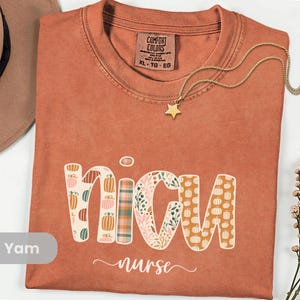 May include: A terracotta-coloured t-shirt with the word "NICU" in a decorative font, featuring pumpkin and floral patterns. The word "nurse" is written below in cursive. A gold necklace with a star pendant is draped across the shirt.