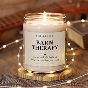 Op de afbeelding: Een brandende kaars in een glazen pot met de tekst "Smells Like Barn Therapy" op een wit etiket. De kaars staat op een ronde houten onderzetter, omringd door lichtslingers. De geur van de kaars roept een gevoel van thuishoren op.