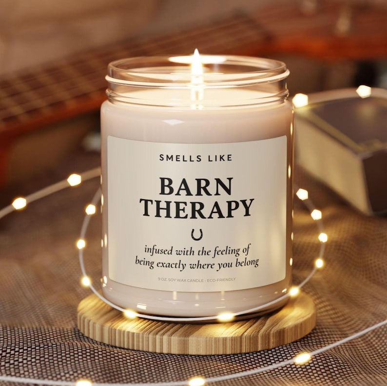 May include: A lit soy wax candle in a glass jar with the text "Smells Like Barn Therapy" on a white label. The candle sits on a round wooden coaster, surrounded by string lights. The candle's scent is described as evoking a sense of belonging.