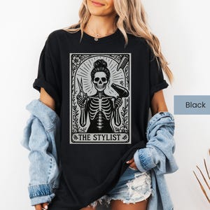 May include: Black t-shirt with a skeleton design holding hairdressing tools, scissors and a hairdryer. The text "THE STYLIST" is displayed within a decorative frame. The shirt is worn by a person.