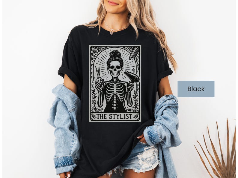 May include: Black t-shirt with a skeleton design holding hairdressing tools, scissors and a hairdryer. The text "THE STYLIST" is displayed within a decorative frame. The shirt is worn by a person.