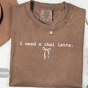 以下が含まれることがあります： 「i need a chai latte.」のテキストとリボンのグラフィックが入ったエスプレッソカラーのTシャツ。ゴールドの星型ペンダントネックレスと茶色の帽子も見える。シャツにはComfort Colorsのタグが付いています。