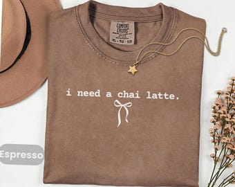 May include: Espresso-colored t-shirt with the text "i need a chai latte." and a bow graphic. A gold star pendant necklace and a brown hat are also visible. The shirt has a Comfort Colors tag.