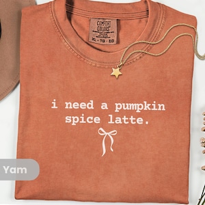 May include: A terracotta-coloured t-shirt with the text "i need a pumpkin spice latte." and a bow graphic. A gold star pendant necklace and a brown hat are also visible. The shirt is labelled XL-TG-EG.