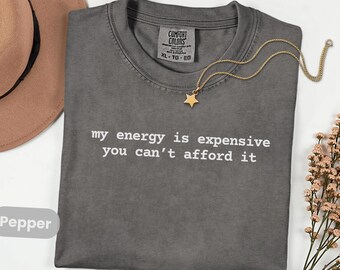 May include: A dark gray t-shirt with the text "my energy is expensive you can't afford it" in white. A gold necklace with a star pendant rests on the shirt. The shirt is labeled "Comfort Colors" and is size XL.