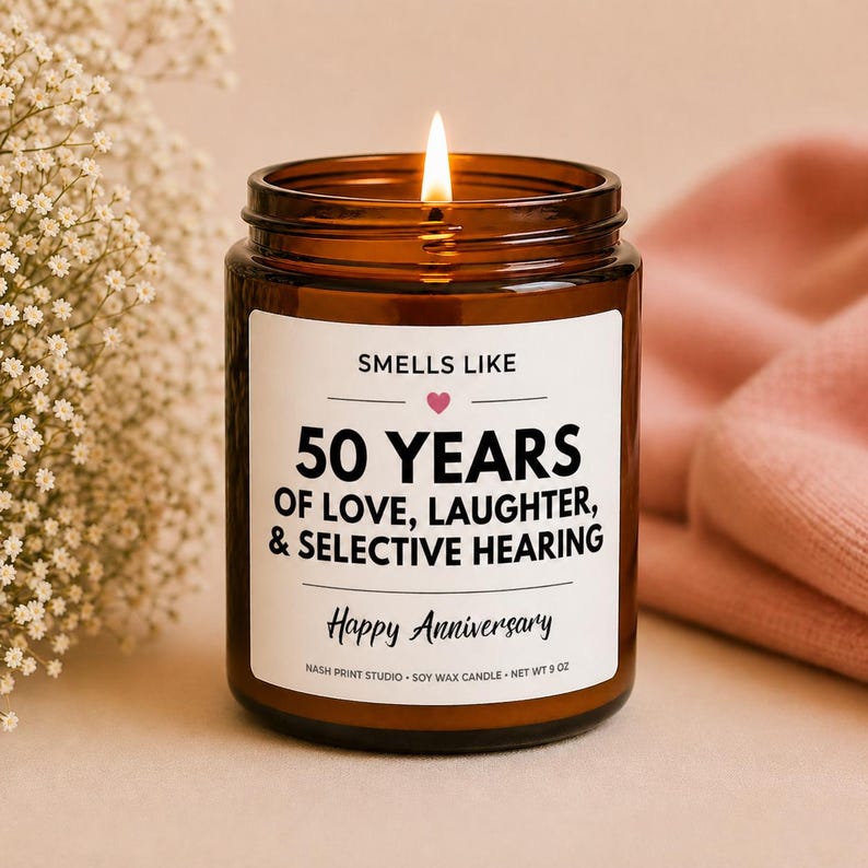 Pode incluir: Uma vela acesa de cera de soja num frasco de vidro castanho. O r&oacute;tulo diz "Smells Like 50 Years of Love, Laughter, & Selective Hearing. Happy Anniversary." A vela pesa 255 gramas.