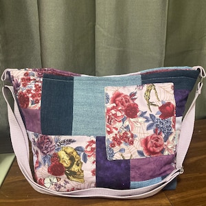 May include: A patchwork shoulder bag with a light purple strap. The bag features a mix of denim and floral fabrics, including a skull and rose design. The bag is a mix of blues, purples, and pinks.