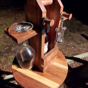 May include: Wooden wine rack with a bottle of wine, a corkscrew, and two wine glasses. The rack is sitting on a wooden table with a round top and three legs.